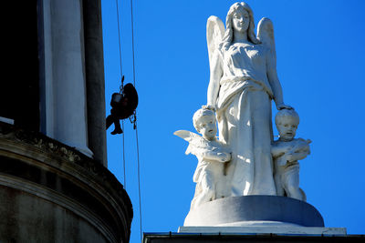 Low angle view of window washer working by angel statue against clear blue sky