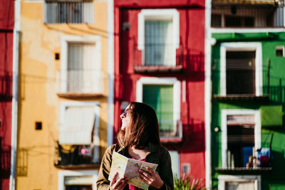 Smiling woman with map standing against wall during sunny day