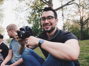 Portrait of man holding camera while sitting by friends at park