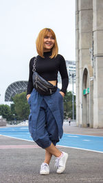 Thoughtful young woman standing on footpath in city against clear sky