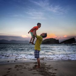 Side view full length of happy father holding son at beach against sky during sunset