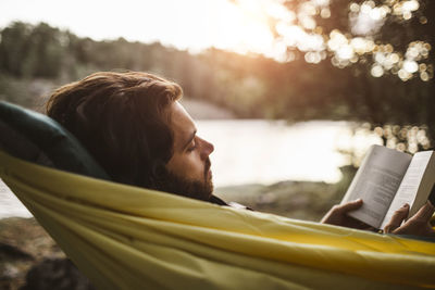 Contemplating man reading book while lying over hammock in forest