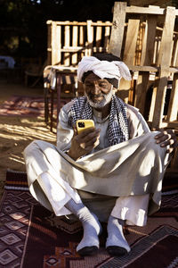 Elderly nubian male in traditional clothes and turban sitting in yard and using mobile phone on sunny day