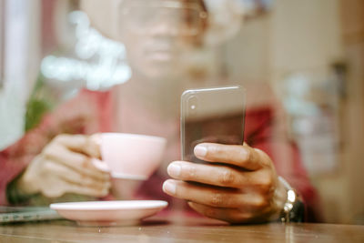 From below stylish african american male in hat sitting at table in coffee shop and browsing smartphone while enjoying fresh hot drink and chilling at weekend