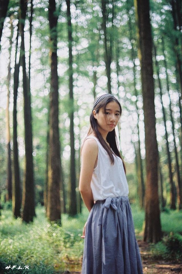 young adult, tree, person, casual clothing, lifestyles, portrait, front view, leisure activity, forest, standing, looking at camera, young women, three quarter length, smiling, focus on foreground, tree trunk, happiness