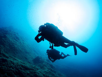 Unrecognizable professional diver in wetsuits and flippers swimming near rough stony formation with uneven surface in deep blue sea with clear water