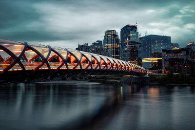 Illuminated bridge over river by buildings against sky in city