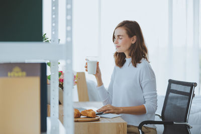 Side view of businesswoman holding milk using laptop on desk in office