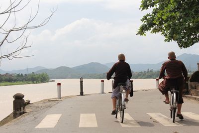 Rear view of friends riding bicycles on road by river against sky