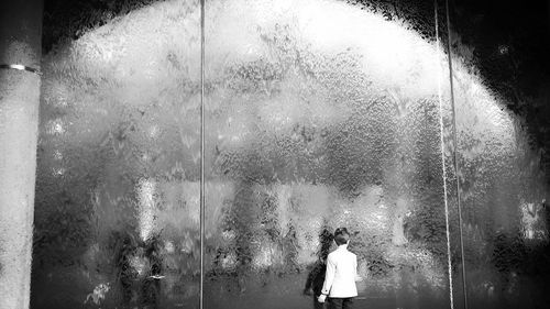 Rear view of boy touching glass wall in city