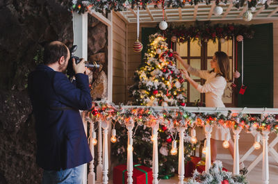 Man photographing woman while decorating christmas tree at home