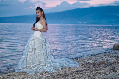 Side view of beautiful bride standing on shore at beach against sky during sunset