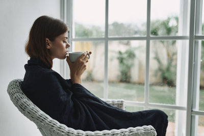 Side view of young woman drinking coffee while sitting by window