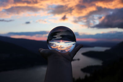 Human hand holding crystal ball against sky during sunset