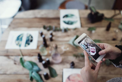 Cropped hands of mid adult female owner photographing brochure and perfume bottles on table at workshop