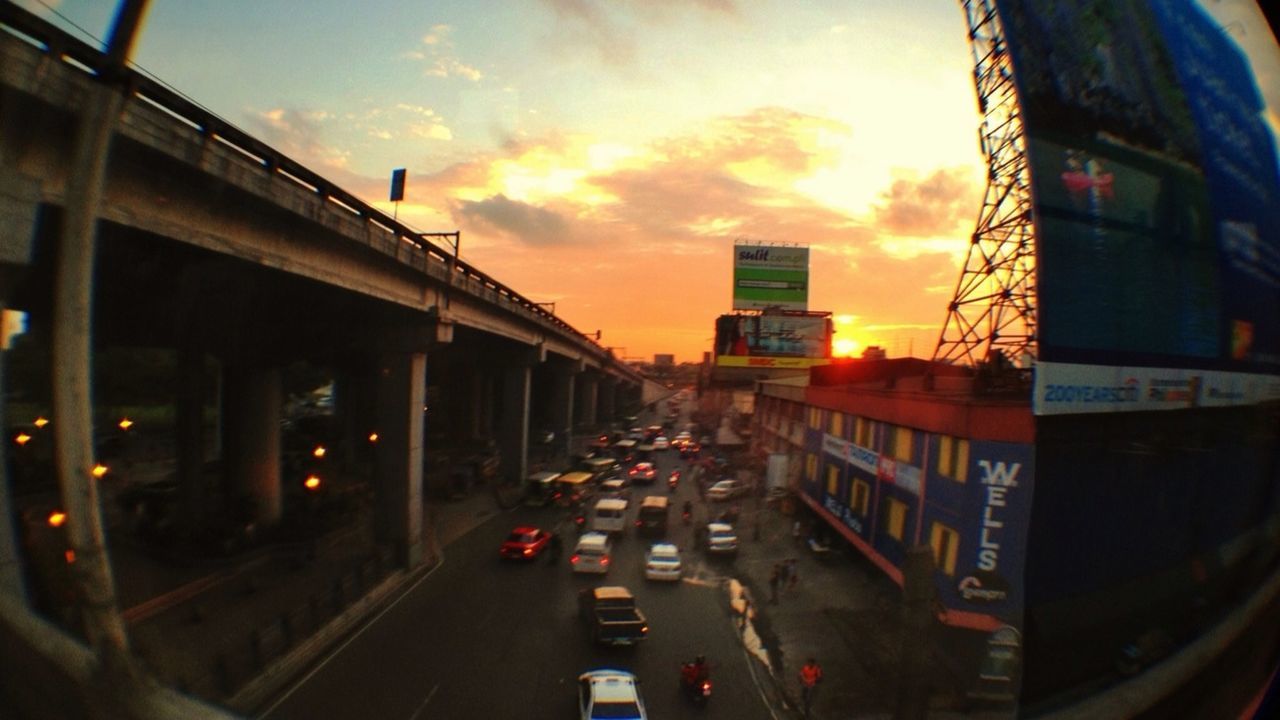 transportation, architecture, built structure, sunset, mode of transport, land vehicle, building exterior, car, city, sky, traffic, railroad track, public transportation, orange color, street, cloud - sky, city life, on the move, road, dusk