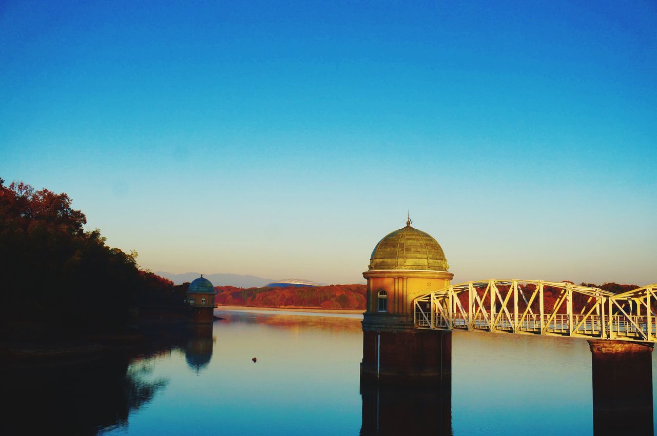 clear sky, water, copy space, blue, built structure, reflection, architecture, waterfront, tranquility, lake, river, tranquil scene, standing water, building exterior, scenics, nature, connection, outdoors, no people, calm