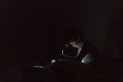 Side view of boy with illuminated headlamp writing homework while sitting on bed in darkroom at home