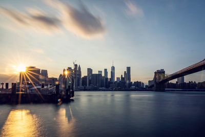 Modern buildings by river against sky during sunset