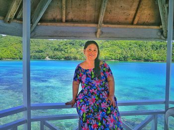 Portrait of smiling woman sitting on railing against sea