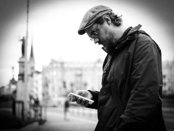 Side view of man using mobile phone while standing against sky in city