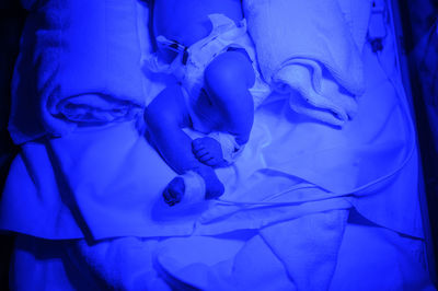 From above crop ill tiny baby lying in infant incubator in neonatal intensive care union under ultraviolet for light therapy in hospital