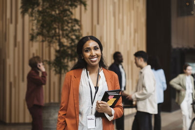 Portrait of smiling female business professional holding diary while standing at convention center