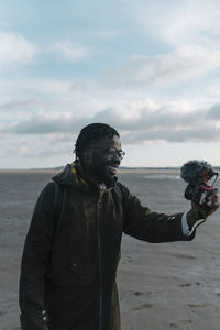 Smiling male vlogger live streaming through camera at beach against sky