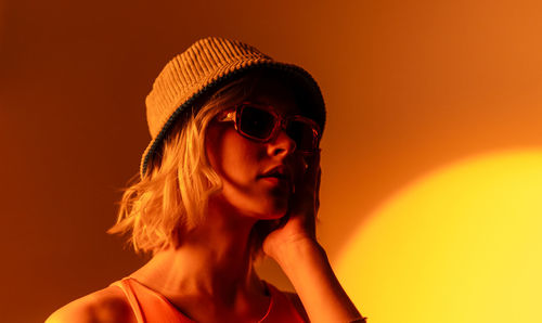 Fashionable model wearing trendy sunglasses and stylish hat against orange background in studio