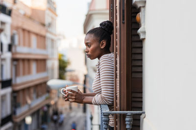 Side view of reflective african american female with cup of hot beverage looking away from balcony in city