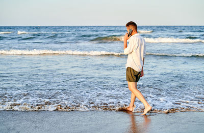 Full body side view of barefoot male manager in white shirt and black shorts walking on beach near sea waves on a phone call on smartphone during summer vacation