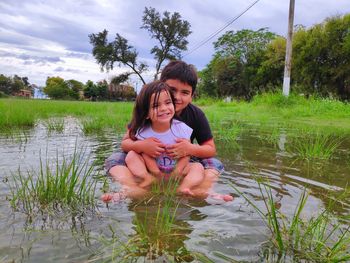 Portrait of brother with cute sister sitting in lake against cloudy sky