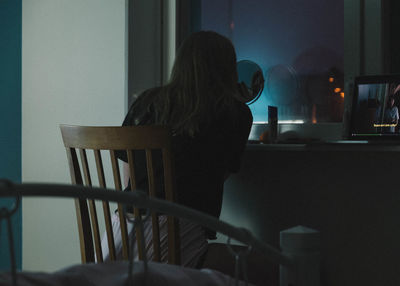 Rear view of woman applying eyeliner while sitting on chair at home