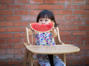 Cute girl holding watermelon while sitting against brick wall