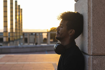 Thoughtful young man with headphones leaning on wall against clear sky during sunset