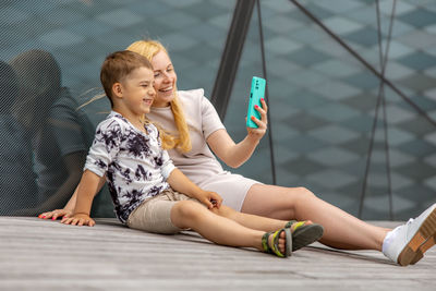 Happy blond woman and little boy sitting on terrace and making selfie on smartphone. mother and son