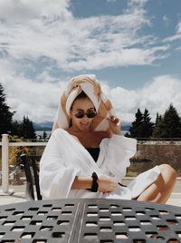 Portrait of woman in towel and sunglasses sitting against sky