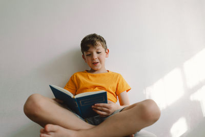 A cute boy wearing an orange t-shirt is sitting on a soft ottoman reading a book