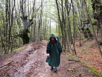 Hiker in raincoat walking under rain on wet clay road in spring forest