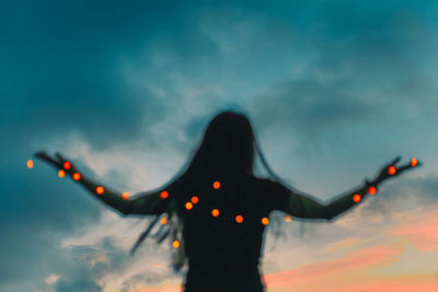 Rear view of silhouette woman with illuminated string light standing by sea against sky