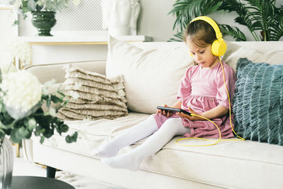 A little girl in yellow headphones sits on a sofa with a phone.