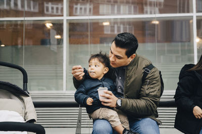 Father feeding baby food to son while sitting at bus stop in city