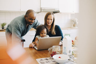 Smiling parents looking at daughter playing with laptop in dining room