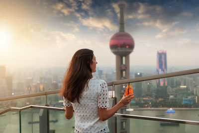 Rear view of mid adult woman looking at cityscape while standing by railing against sky during sunset