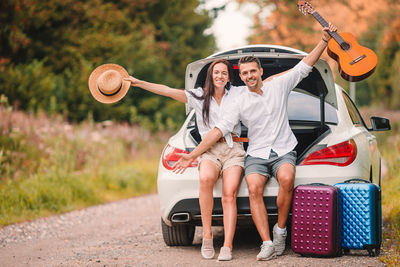 Portrait of couple holding hat and guitar sitting on car trunk