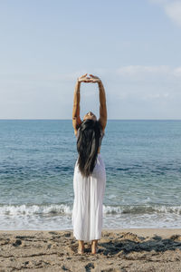 Back view of ethnic female leaning back while standing in ashta chandrasana pose during yoga practice on sandy coast against ocean