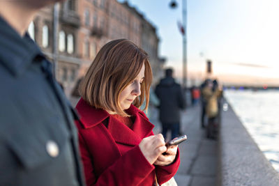 Young woman with red hair, on the embankment of the river in the city, typing text on a smartphone. 