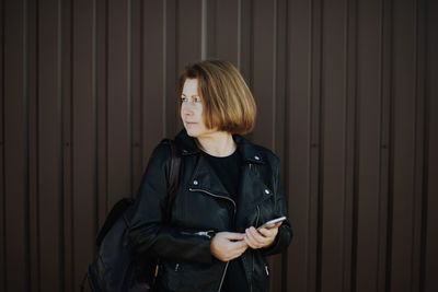 Young woman using phone while standing against wall