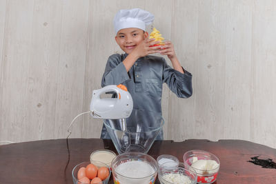 Portrait of boy wearing chef hat with ingredients on table standing against wall at home