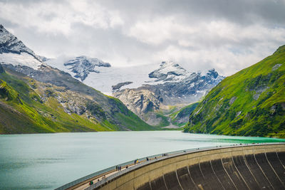 Mountains peak covered in snow and grass on a cloudy day. a big water reservoir and a dam.